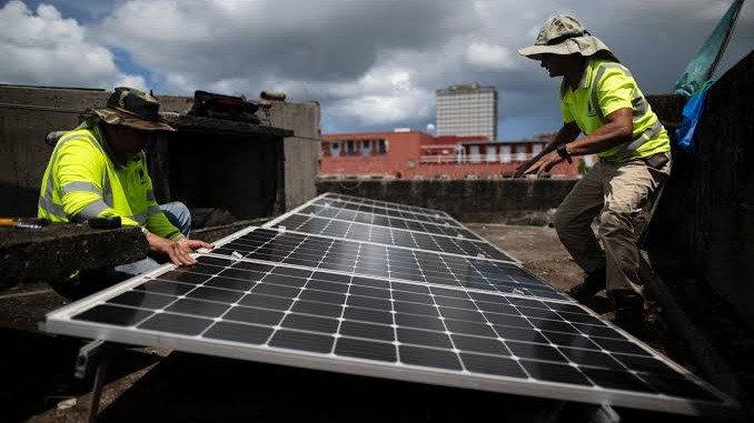 Dos técnicos con chalecos reflectantes amarillos instalan paneles solares en la azotea de un edificio, con nubes y edificios al fondo