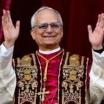 El papa León XIV, con estola bordada y muceta roja, saluda con ambos brazos en alto ante una cortina roja