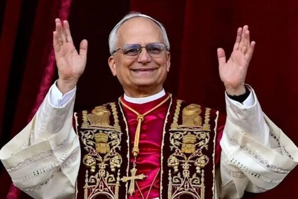 El papa León XIV, con estola bordada y muceta roja, saluda con ambos brazos en alto ante una cortina roja
