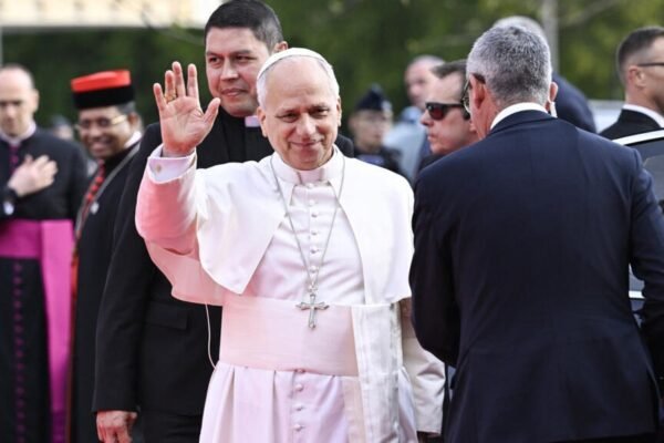 El papa León XIV, vestido de blanco, saluda con la mano y sonríe rodeado de clérigos y miembros de seguridad junto a un vehículo oficial