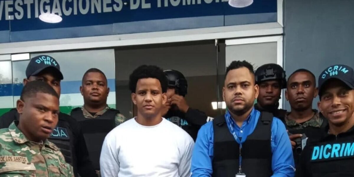 Bryan Rafael Gómez, de ropa blanca, custodiado por agentes de la DICRIM, militares y un representante del Ministerio Público frente al letrero de Investigaciones de Homicidios