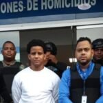 Bryan Rafael Gómez, de ropa blanca, custodiado por agentes de la DICRIM, militares y un representante del Ministerio Público frente al letrero de Investigaciones de Homicidios
