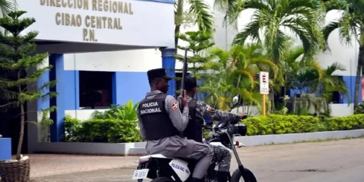 Dos agentes de la Policía Nacional en una motocicleta pasan frente a la sede de la Dirección Regional Cibao Central; el que va atrás porta un arma larga.