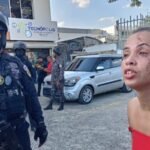 Agentes de la Policía Nacional con casco y armas largas junto a una cinta de acordonamiento; en primer plano, una mujer de blusa roja con raspaduras en el rostro habla con gesto de angustia