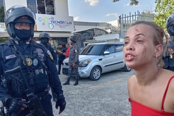 Agentes de la Policía Nacional con casco y armas largas junto a una cinta de acordonamiento; en primer plano, una mujer de blusa roja con raspaduras en el rostro habla con gesto de angustia