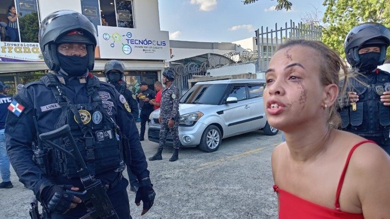 Agentes de la Policía Nacional con casco y armas largas junto a una cinta de acordonamiento; en primer plano, una mujer de blusa roja con raspaduras en el rostro habla con gesto de angustia