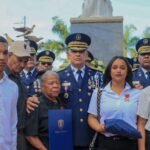 La Policía rinde honores a cuatro agentes caídos en servicio Un alto oficial de la Policía Nacional con uniforme de gala abraza a familiares de agentes caídos, que sostienen una bandera doblada y un diploma, frente a un monumento