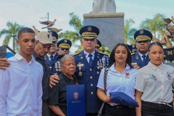 Un alto oficial de la Policía Nacional con uniforme de gala abraza a familiares de agentes caídos, que sostienen una bandera doblada y un diploma, frente a un monumento