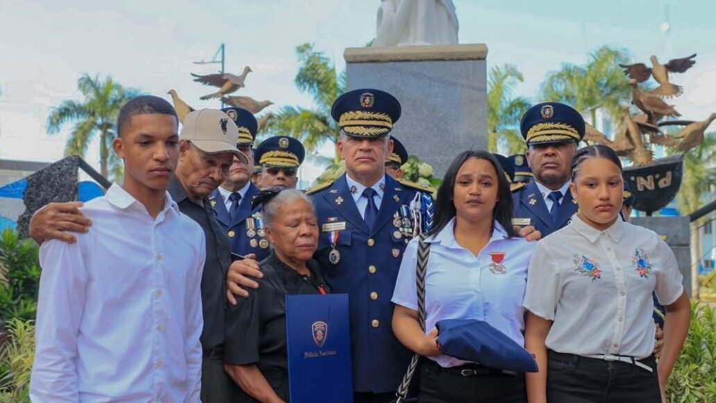 Un alto oficial de la Policía Nacional con uniforme de gala abraza a familiares de agentes caídos, que sostienen una bandera doblada y un diploma, frente a un monumento