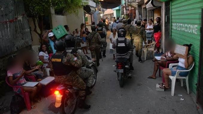 Agentes de la Policía Nacional y militares en motocicleta y a pie recorren un callejón estrecho mientras vecinos observan sentados frente a sus casas