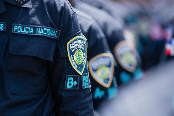 Detalle de la manga del uniforme de la Policía Nacional con el escudo institucional, con agentes en fila al fondo.