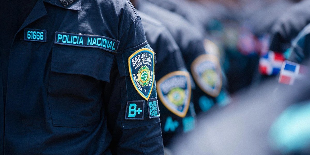 Detalle de la manga del uniforme de la Policía Nacional con el escudo institucional, con agentes en fila al fondo.