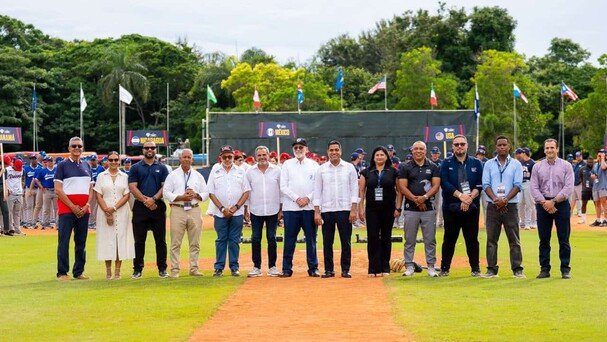 Grupo de dirigentes y autoridades de pie sobre un terreno de béisbol, con banderas de varios países y jugadores al fondo
