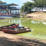 Fuerza del Pueblo denuncia irregularidades en la presa de Hatillo Botes de madera varados en la orilla de un embalse de aguas verdosas, con una embarcación turística de dos pisos y vehículos estacionados al fondo.
