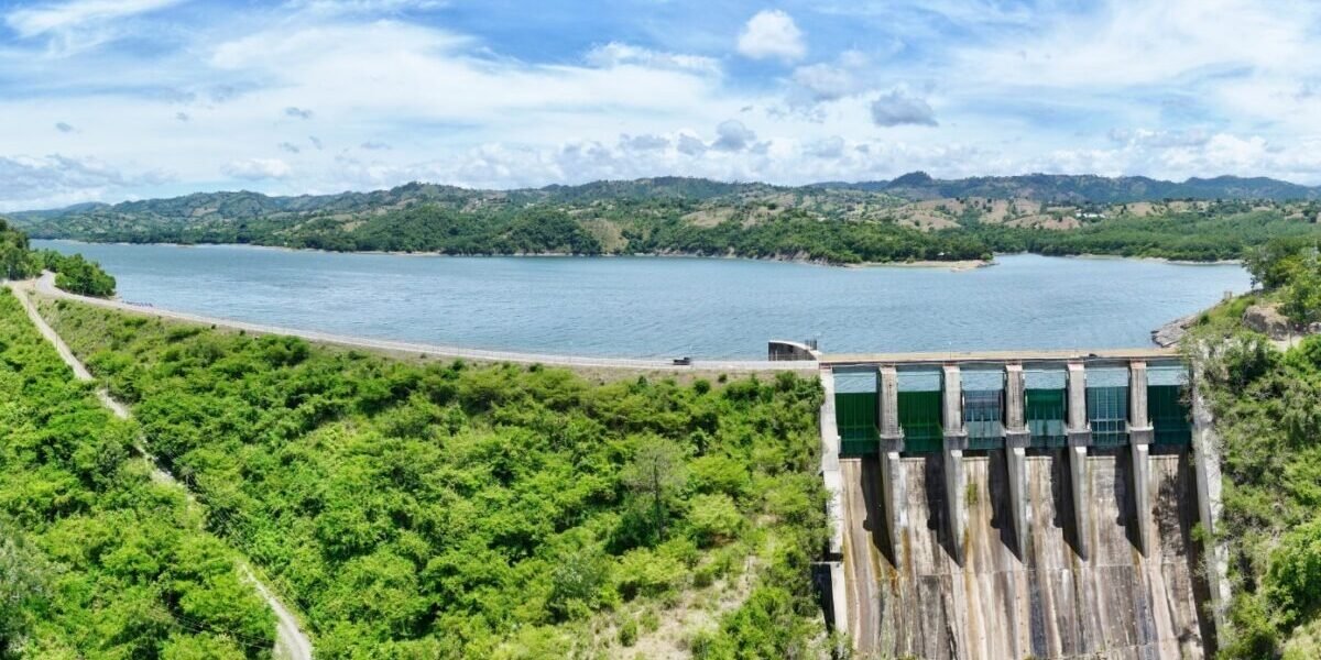 Vista aérea de una presa con su vertedero de compuertas verdes a la derecha y un embalse amplio rodeado de colinas con vegetación bajo un cielo con nubes.