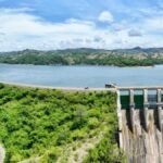 Las presas suben al 62 % de su capacidad gracias a las lluvias de octubre Vista aérea de una presa con su vertedero de compuertas verdes a la derecha y un embalse amplio rodeado de colinas con vegetación bajo un cielo con nubes.