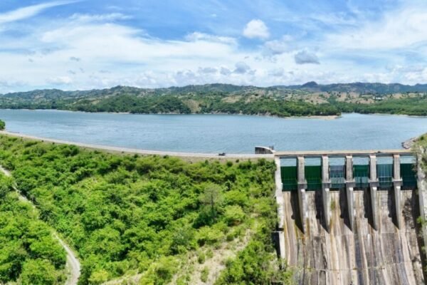Vista aérea de una presa con su vertedero de compuertas verdes a la derecha y un embalse amplio rodeado de colinas con vegetación bajo un cielo con nubes.