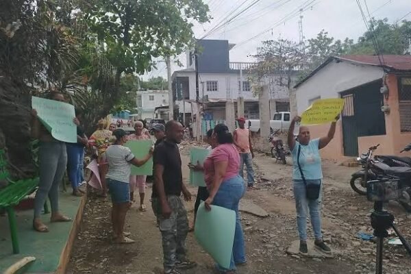 Vecinos con carteles de papel en una calle de tierra con piedras, casas y motocicletas al fondo