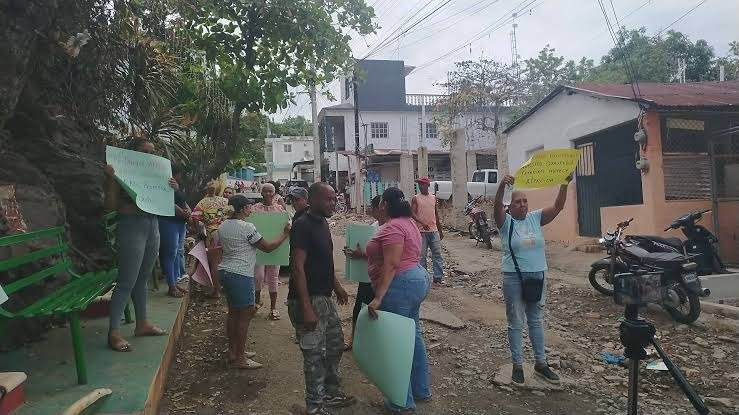 Vecinos con carteles de papel en una calle de tierra con piedras, casas y motocicletas al fondo