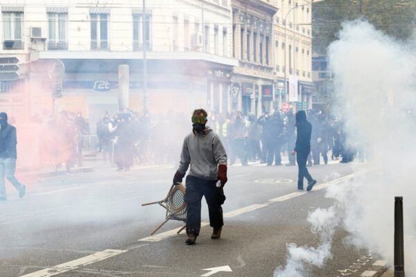 Un manifestante con máscara antigás y gafas arrastra una silla por una calle llena de humo; al fondo, personas encapuchadas