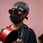 Una manifestante con gafas oscuras, mascarilla negra y una cinta en la frente contra LUMA golpea un caldero con una cuchara
