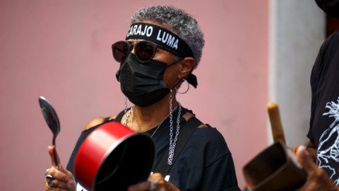 Una manifestante con gafas oscuras, mascarilla negra y una cinta en la frente contra LUMA golpea un caldero con una cuchara