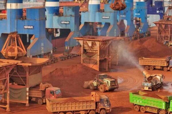 Grúas portuarias, cargadoras y camiones mueven montones de mineral rojizo en un muelle de carga