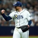 Un lanzador de los Rays de Tampa Bay, con barba y gorra azul, levanta el puño y grita de celebración en el terreno con el público al fondo