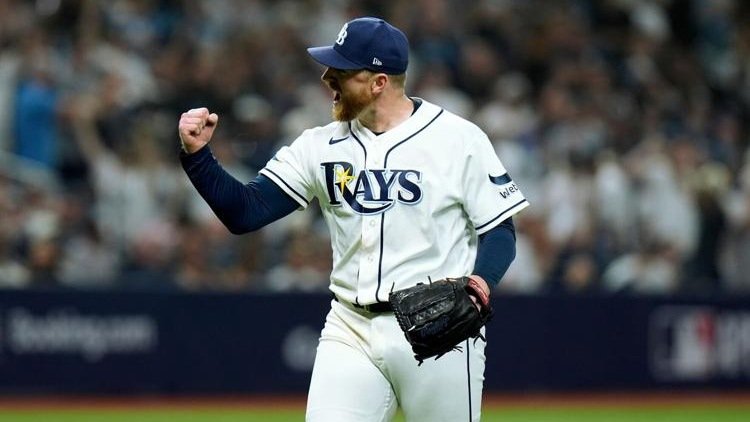 Un lanzador de los Rays de Tampa Bay, con barba y gorra azul, levanta el puño y grita de celebración en el terreno con el público al fondo