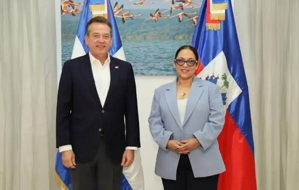 Un hombre de chaqueta oscura y una mujer de chaqueta gris claro posan de pie entre una bandera dominicana y una bandera haitiana, frente a un cuadro de flamencos