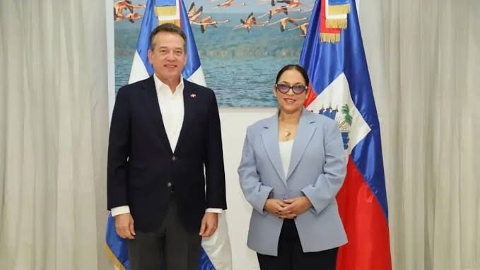 Un hombre de chaqueta oscura y una mujer de chaqueta gris claro posan de pie entre una bandera dominicana y una bandera haitiana, frente a un cuadro de flamencos