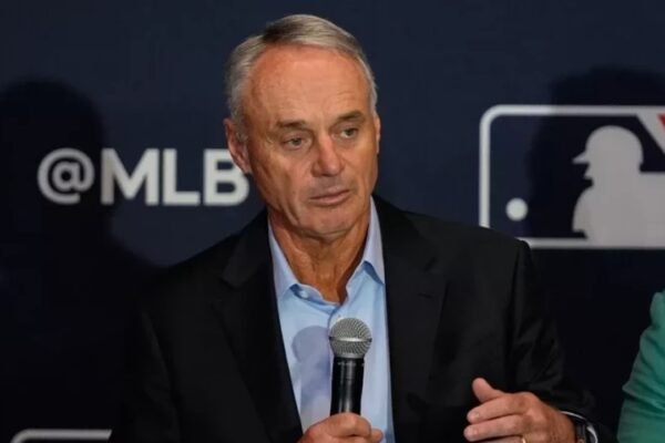 El comisionado de las Grandes Ligas, Rob Manfred, habla con un micrófono en una rueda de prensa ante el logo de MLB