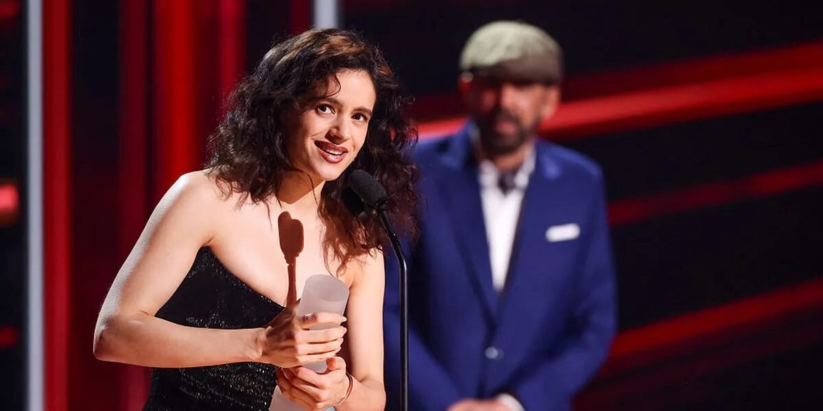 Rosalía sonríe frente al micrófono con un trofeo en las manos mientras Juan Luis Guerra, con gorra y traje azul, la observa al fondo del escenario