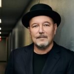 Rubén Blades, con sombrero negro y saco oscuro de rayas, posa apoyado en una pared en un pasillo