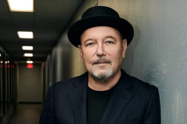 Rubén Blades, con sombrero negro y saco oscuro de rayas, posa apoyado en una pared en un pasillo