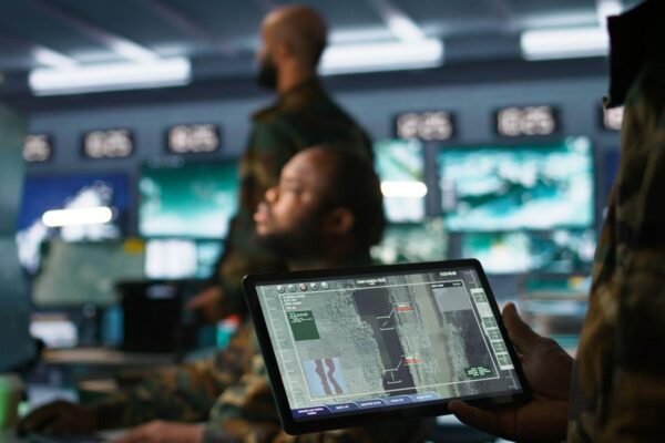 Militares trabajan en una sala de control con pantallas; uno sostiene una tableta con imágenes de vigilancia aérea