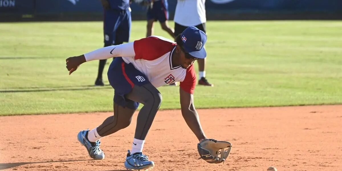 Un jugador con uniforme y gorra de la selección dominicana se agacha para atrapar con el guante una pelota rodada en el cuadro; detrás, otros miembros del equipo observan.