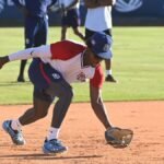 Un jugador con uniforme y gorra de la selección dominicana se agacha para atrapar con el guante una pelota rodada en el cuadro; detrás, otros miembros del equipo observan.