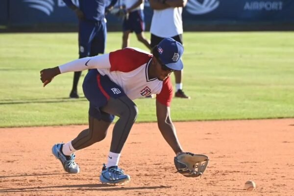 Un jugador con uniforme y gorra de la selección dominicana se agacha para atrapar con el guante una pelota rodada en el cuadro; detrás, otros miembros del equipo observan.