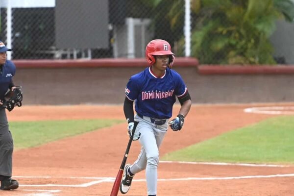 Bateador de la selección dominicana con casco rojo y uniforme azul corre tras batear; detrás, el árbitro de home
