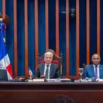 Integrantes de la mesa directiva del Senado dominicano sentados en el estrado durante una sesión, entre dos banderas dominicanas