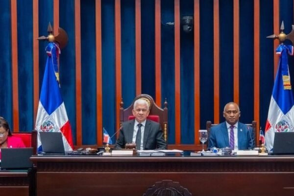 Integrantes de la mesa directiva del Senado dominicano sentados en el estrado durante una sesión, entre dos banderas dominicanas