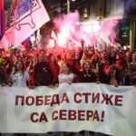 Jóvenes marchan de noche por una calle con banderas serbias y bengalas detrás de una pancarta blanca con letras rojas en cirílico