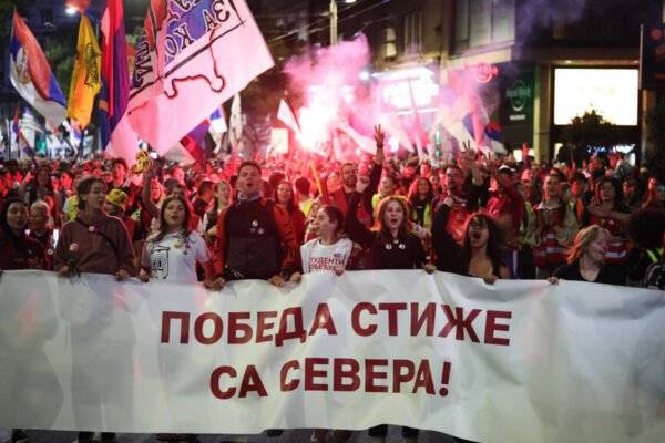 Jóvenes marchan de noche por una calle con banderas serbias y bengalas detrás de una pancarta blanca con letras rojas en cirílico