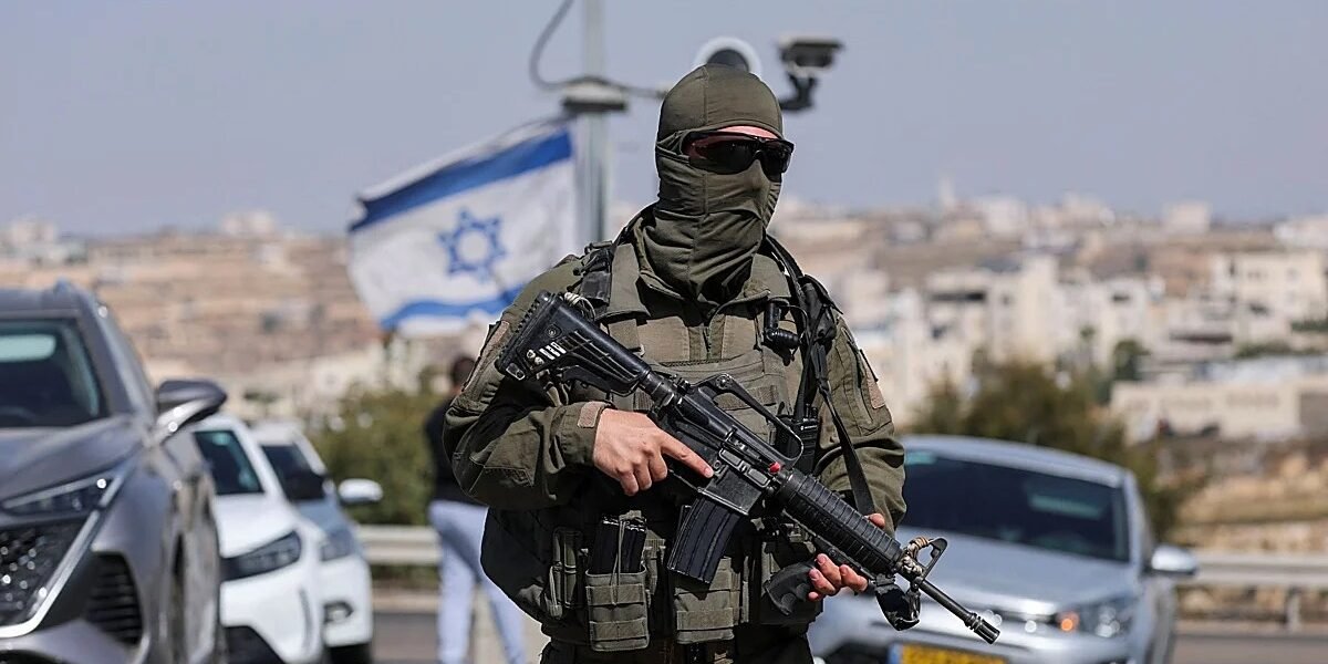 Militar israelí con el rostro cubierto y un fusil junto a una carretera con autos, una bandera de Israel y una ciudad al fondo