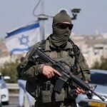 Militar israelí con el rostro cubierto y un fusil junto a una carretera con autos, una bandera de Israel y una ciudad al fondo