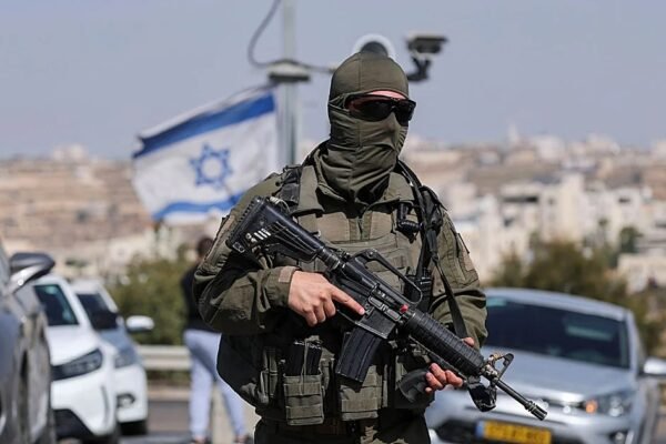 Militar israelí con el rostro cubierto y un fusil junto a una carretera con autos, una bandera de Israel y una ciudad al fondo