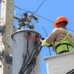 Un técnico con casco naranja y chaleco reflectante trabaja desde una canasta elevada en los transformadores de un poste eléctrico