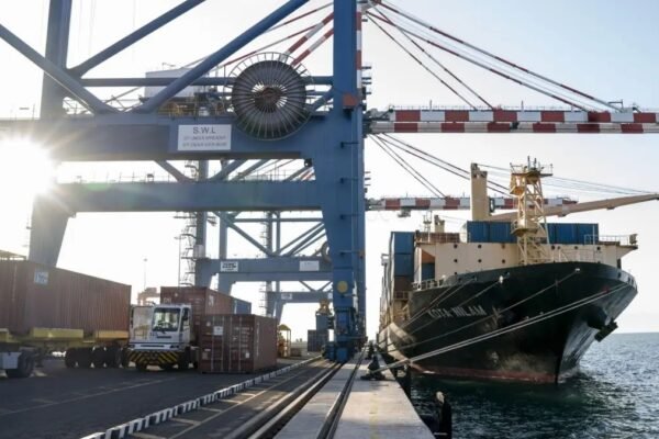 Grúas pórtico azules sobre un muelle con camiones y contenedores, junto a un buque portacontenedores atracado