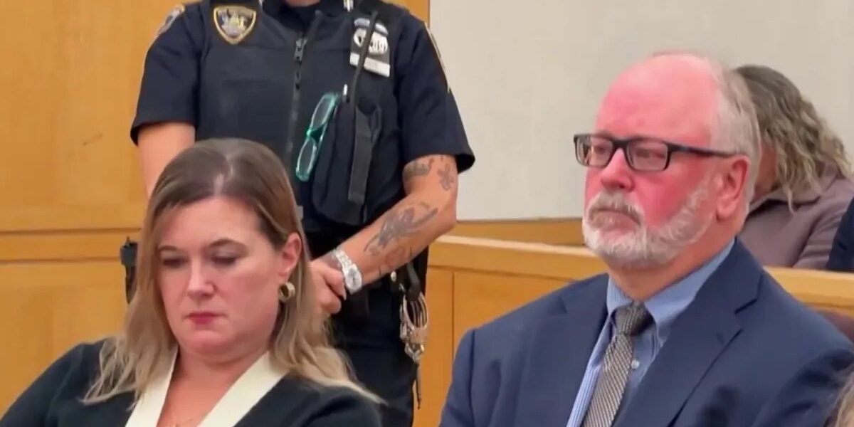 Una mujer de cabello rubio, vestida de negro, sentada con la mirada baja en una sala de tribunal; a su lado, un hombre de traje y lentes, y detrás, una agente uniformada